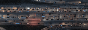 Aerial view of a congested multi-lane highway at dusk, with dense vehicle traffic and glowing headlights stretching across an urban freeway interchange.